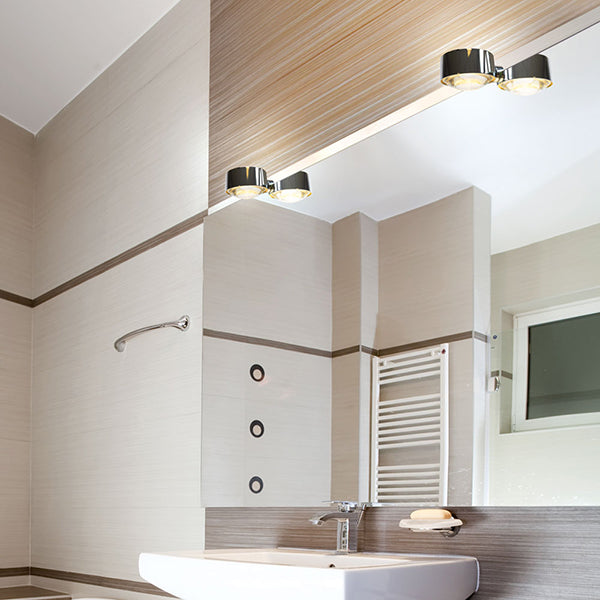 Ein modernes Badezimmer mit hellen Wänden und chromfarbenen Akzenten. Ein großer Spiegel wird von einer chromfarbenen Leuchte erhellt.