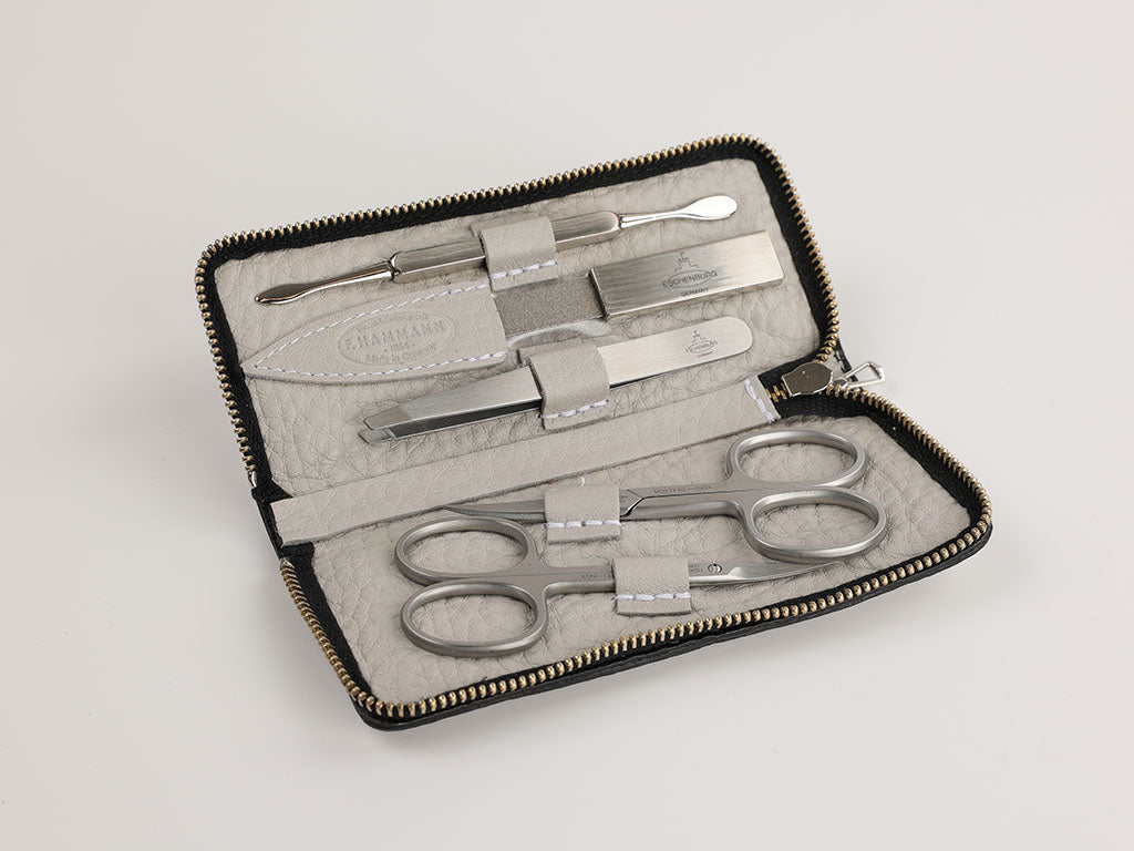 Ein geöffnetes Maniküre-Set in einem grauen Etui mit Reißverschluss. Das Set enthält mehrere Werkzeuge, darunter Scheren, eine Nagelfeile, eine Pinzette und einen Nagelhautschieber, alle ordentlich in Halterungen befestigt.