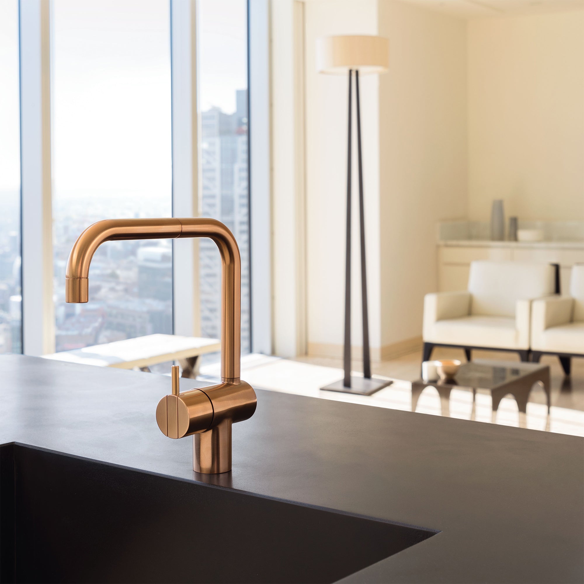 Ein moderner Wasserhahn in gebürstetem Kupfer von Vola KV1, montiert auf einer schwarzen Küchentheke mit Blick auf ein elegantes, lichtdurchflutetes Wohnzimmer im Hintergrund. Erhältlich bei uns auf dasfeinebad.