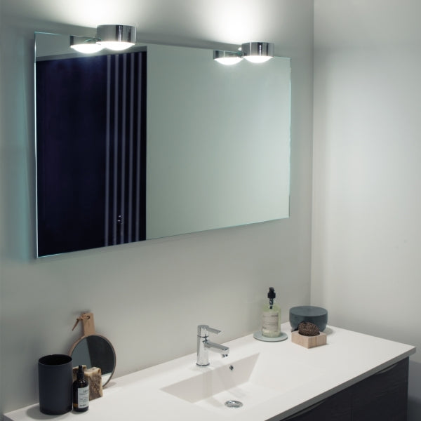 Modernes Badezimmer mit weißen Fliesen und chromfarbenen Akzenten. Der große Spiegel wird von einer chromfarbenen Leuchte beleuchtet.